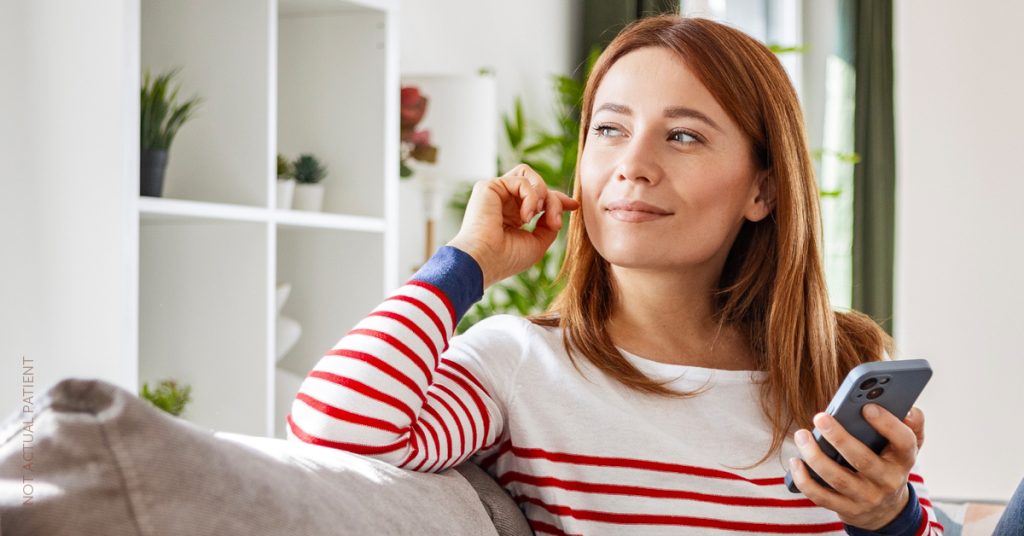 Woman sitting on a couch looking thoughtful and holding a phone (model)